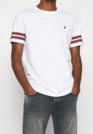 Valkoinen puuvillainen t-paita, jossa on lyhyet hihat, viininpunaiset raidat hihoissa ja pieni logo rinnassa. Rentoa istuvuutta ja pyöreä pääntie.