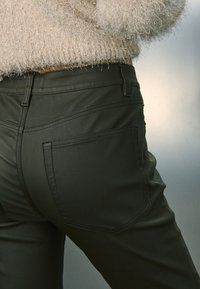 Maslinasto zelene traperice glatkoga tekstura, s klasičnim dizajnom s pet džepova i zaobljenim stražnjim džepovima, uparene s mekanim svijetlim džemperom.