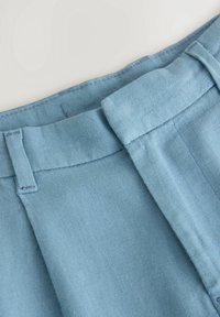 Ceinture de pantalon en lin bleu clair avec passants pour ceinture et fermeture frontale dissimulée, présentée sur un fond blanc.