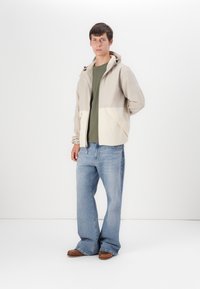 Lichtbeige hoodie-jack met crèmekleurige zakken, gecombineerd met een groene shirt en blauwe wijde jeans. Bruine schoenen maken de outfit af.