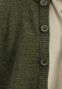 Olive green knitted cardigan with a ribbed collar and five gray buttons along the front. Underneath, a light beige top is visible.