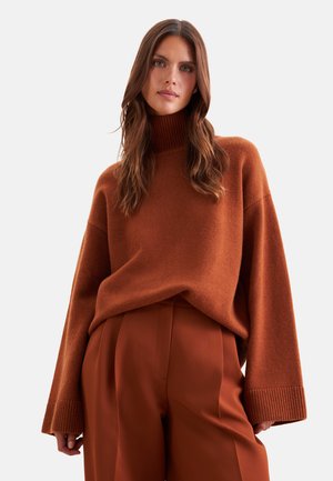Femme aux longs cheveux bruns portant un pull à col roulé marron et un pantalon large taille haute assorti, debout devant un fond uni.