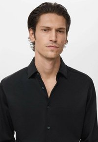 Hombre joven con el cabello oscuro peinado hacia atrás, vistiendo una camisa negra de botones, mirando hacia delante con una expresión neutral, sobre un fondo blanco.