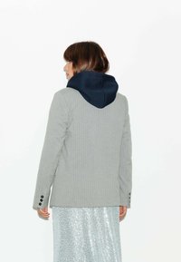Blazer gris à rayures avec une capuche bleu marine, présentant une coupe cintrée et des poignets boutonnés. Porté par-dessus une jupe en sequins argentés scintillants.