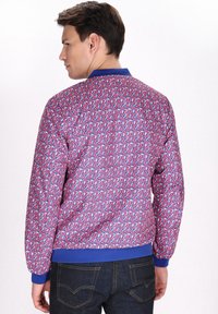 Giacca bomber colorata con un motivo astratto ripetuto in rosso, blu e bianco. Presenta un colletto e polsini a costine blu.
