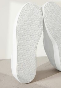 Semelles de baskets blanches avec un motif de grip texturé et le logo "NEXT" en relief. Fini lisse, bords arrondis et marquages de taille visibles.