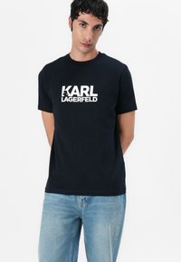 Marineblå t-skjorte med kort erm laget av bomull, med et fremtredende hvitt trykk av "KARL LAGERFELD" på forsiden. Klassisk rund hals.
