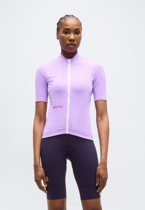 Kobieta ubrana w krótkim rękawem lawendowy strój kolarski z zamkiem błyskawicznym z przodu oraz czarne, wyściełane szorty kolarskie, stojąca na jednolitym tle.