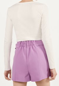 Femme portant un short taille haute lilas avec une ceinture froncée et un haut court à manches longues écru, vue de dos.