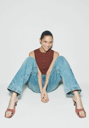 Kvinde med håret bundet tilbage iført en ærmeløs brun top, blå jeans og lyserøde sandaler med hæl, sidder på gulvet og smiler med hænderne foldet.