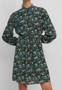Vestido floral con un fondo negro, que presenta patrones en azul, verde y marrón. Manga larga, cintura fruncida y un cuello alto.
