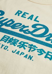 Kremowa bawełniana koszulka z teksturowanym turkusowym nadrukiem z napisem "REAL Super Dry" i japońskimi znakami na dole.