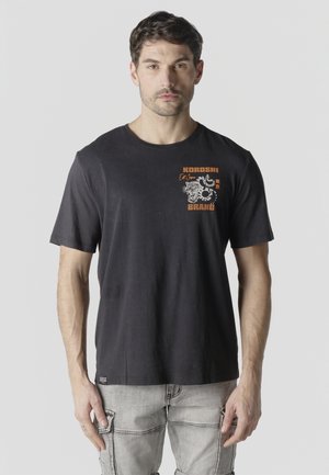 Man draagt een zwarte T-shirt met een oranje-witte afbeelding en tekst op de linker bovenborst, gecombineerd met lichtgrijze cargobroek.