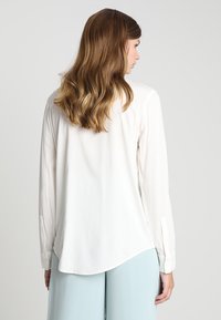 Camicia bianca a maniche lunghe con orlo curvo e polsini con bottoni, caratterizzata da una texture liscia e una caduta fluida in una vestibilità comoda.