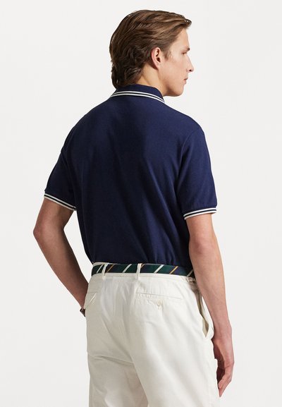 Navy polo shirt with white accents, short sleeves, and a collar. Worn with white trousers and a multicolored striped belt.