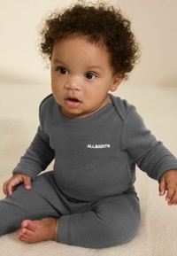 Graues Baby-Strampler mit Waffelstruktur, langen Ärmeln und rundem Hals. Mit dem weißen "ALLSAINTS"-Logo auf der Brust. Das Baby sitzt auf einer weichen Unterlage.