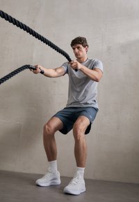 Gray athletic t-shirt and blue shorts worn by a man using black battle ropes, set against a concrete wall and gray floor. White sneakers.