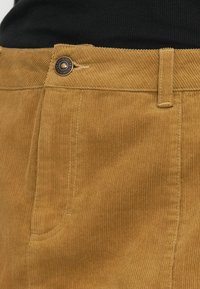 Παντελόνια από κορδυρό σε ταμπά απόχρωση με κάθετη ραβδωτή υφή. Διαθέτουν κούμπωμα με κουμπί, θηλιές ζώνης και λεπτομέρειες εμπρός τσέπης.