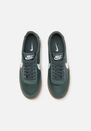 Een paar donker groene Nike-sneakers met witte swoosh-logo's, zwarte veters en bruine rubberen zolen, gezien van bovenaf op een witte achtergrond.