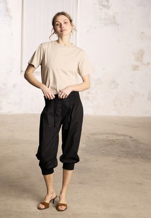 Vrouw staat binnen, draagt een beige shirt met korte mouwen, zwarte cropped broek met elastische manchetten en bruine sandalen met hak.