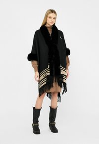 Donna che indossa un poncho nero con frange e rifiniture in pelliccia sopra un vestito marrone e stivali neri con lacci a occhiello, in piedi su uno sfondo bianco.