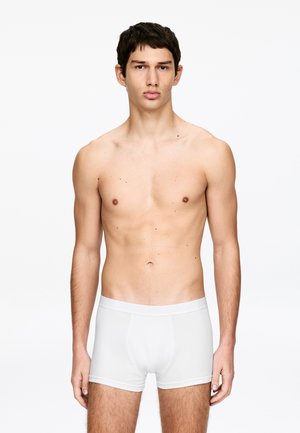 Jeune homme debout devant un fond blanc, portant uniquement un boxer blanc, regardant vers l’avant avec une expression neutre.