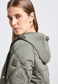 Veste rembourrée vert olive avec un motif texturé et matelassé. Dotée d'une capuche avec cordons de serrage et d'une patch logo sur la manche.