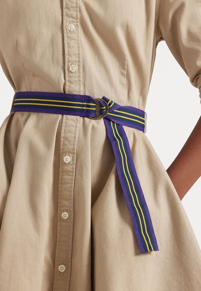 Violets josta ar dzeltenām un zaļām svītrām, ar metāla gredzenu sprādzi, uzvilkta virs bēša krekliņu kleitas ar pogu detaļām.