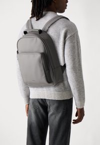 Mochila gris hecha de material suave, con correas ajustables, bolsillo frontal y diseño minimalista. La lleva puesta una persona con un suéter de punto.