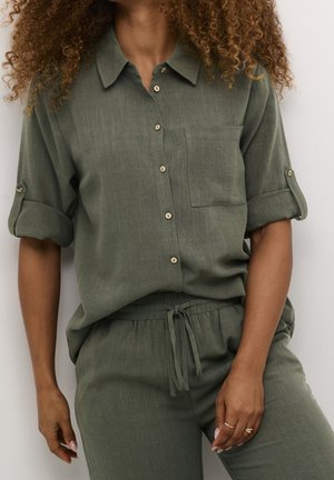 Femme aux cheveux bouclés portant une chemise verte à boutons avec manches retroussées et un pantalon à cordon assorti, debout devant un fond clair.