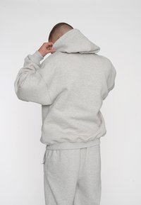Hellgrauer Hoodie mit großer Kapuze, gerippten Bündchen und abgerundetem Saum. Weiche Textur, rückwärts getragen mit lockerer Passform und passender Hose.