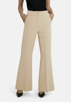 Frau trägt beige hochtaillierte, weit geschnittene Hosen mit vorderen Falten, Seitentaschen und Knopfverschluss, kombiniert mit schwarzen spitz zulaufenden Schuhen und schwarzem Oberteil.