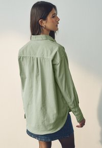 Chemise oversized vert clair avec col à boutons, plis dans le dos et manches longues, associée à une jupe en denim bleue.