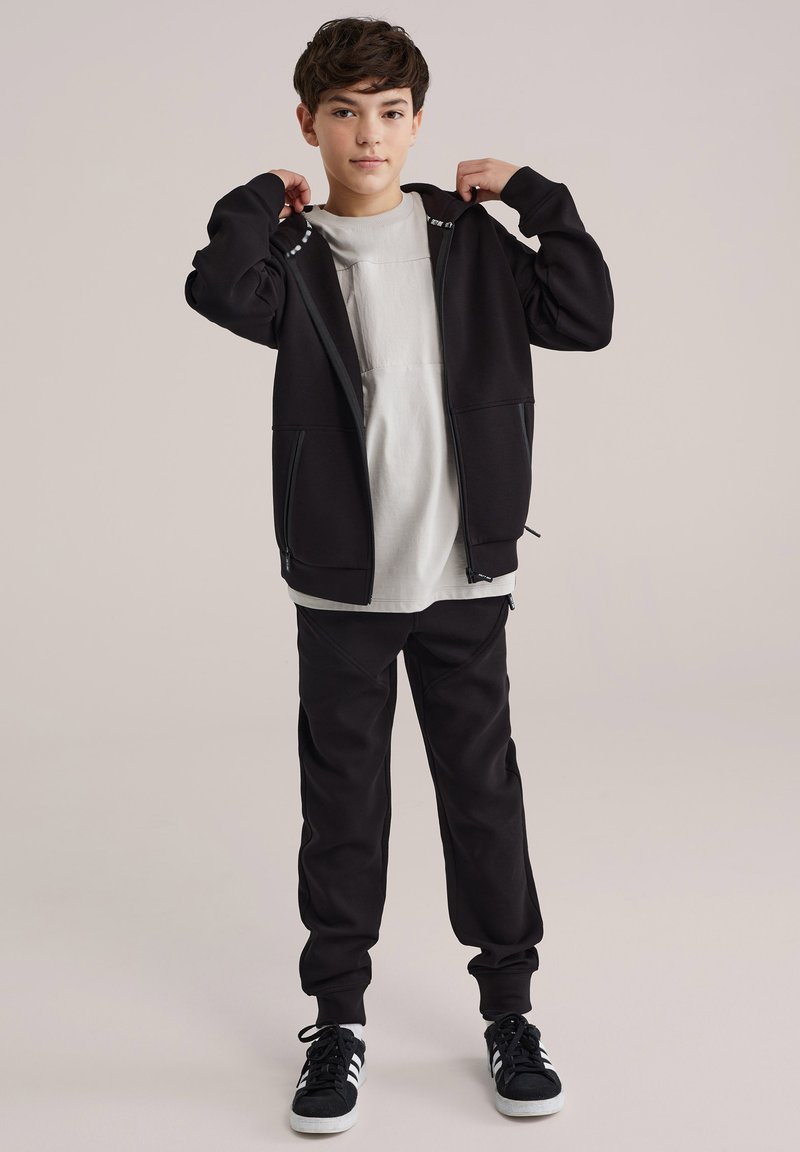 Sweat à capuche zippé noir avec poches, superposé à un t-shirt gris clair, accompagné d'un pantalon de survêtement noir et de baskets noires à rayures blanches.