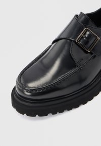 Μαύρο δερμάτινο παπούτσι με στρογγυλεμένη τετράγωνη μύτη, διακοσμητικό ράψιμο, λουράκι με ασημένια αγκράφα και παχύ μαύρο καουτσούκ σόλα με ανάγλυφο πέλμα.