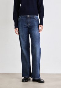 Jeans de denim azul de pierna ancha con un ligero desvanecimiento, diseño de cinco bolsillos y herrajes de cobre. Combinados con un suéter de punto oscuro.