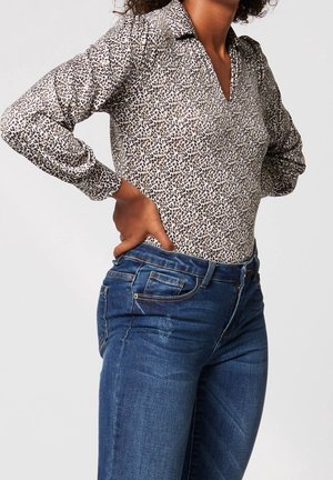 Kvinde iført en langærmet leopardprintbluse stoppet ned i blå denimjeans med hænderne hvilende på hofterne mod en ensfarvet baggrund.