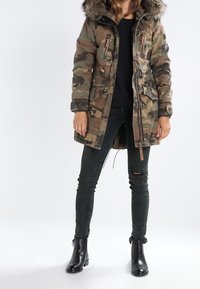 Kobieta w kamuflażowej parkowej kurtce z futrzanym kapturem, czarnych przetartych rurkach, czarnych botkach do kostek, stojąca na tle jednolitego tła.