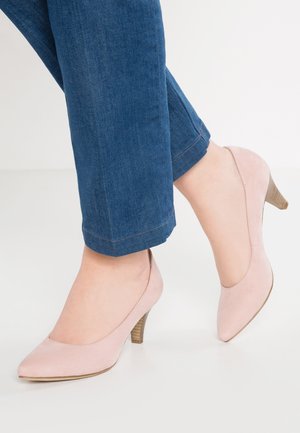 Jasnoróżowe zamszowe buty na wysokim obcasie z zaostrzonym noskiem i drewnianym obcasem, zestawione z podwiniętymi niebieskimi dżinsami.