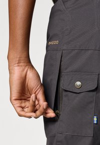 Grå G-1000 tygbyxor med blixtlåsficka, tryckknappstängning och en liten svensk flagga på sidan. Texturerad yta synlig.