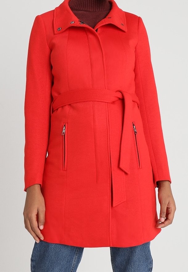Manteau en laine rouge longueur genou avec ceinture nouée à la taille, col montant, poches latérales zippées, porté sur un col roulé bordeaux et un jean bleu.