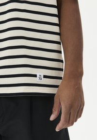 Smėlinio atspalvio marškiniai su juodomis horizontaliomis juostelėmis, turintys mažą etikėtę su užrašu "M 73," pagaminti iš minkštos medvilnės medžiagos.