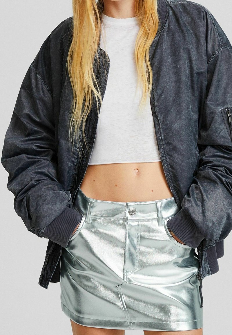 Veste bomber noire avec une texture usée, assortie à une mini-jupe argentée métallique brillante avec des poches et une fermeture à boutons sur le devant.
