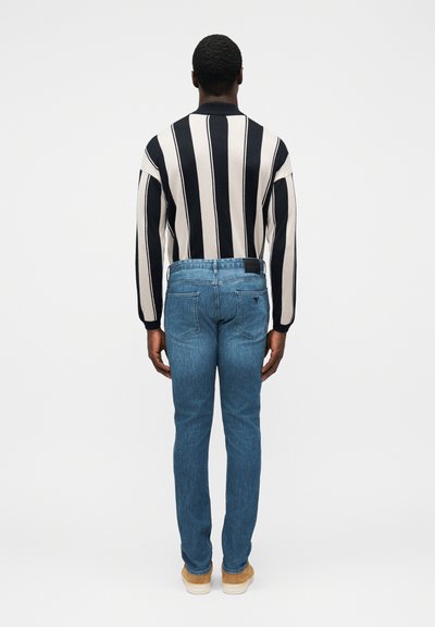 Črtast črno-krem pulover, modre kavbojke z zadnjimi žepi, obute v svetlo rjave čevlje. Model viden od zadaj na enobarvnem ozadju.