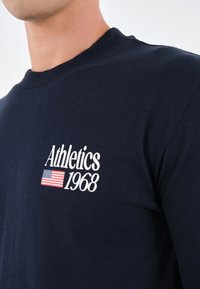 Marineblaue Baumwoll-Hemd mit langen Ärmeln, das weißen Text "Athletics 1968" und ein kleines Grafik der amerikanischen Flagge zeigt. Gerippter Kragen.