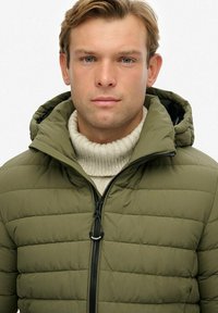 Oliwkowa kurtka puchowa z kapturem, z suwakiem z przodu i kremowym, prążkowanym swetrem pod spodem. Kurtka ma pikowaną teksturę.