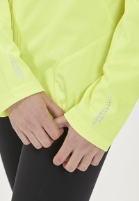 Högsynlig neongul jacka med en slät textur, med reflektiv prickig detaljering på ärmarna och en åtsittande design.