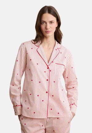 Vrouw die witte en rood gestreepte pyjama draagt met kleine rode hartjespatronen, staand met handen in de zakken tegen een effen achtergrond.