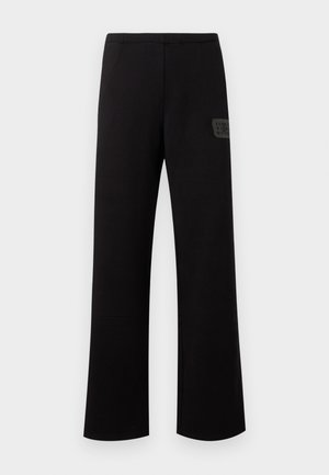 Homme debout portant un sweat à capuche noir avec un logo sur le devant, un pantalon noir ample et des chaussures noires sur un fond gris uni.