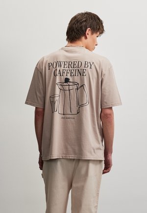 Jeune homme de dos portant un t-shirt beige avec le contour noir d'une cafetière, d'une tasse, et le texte « Powered by Caffeine and Ambition. »